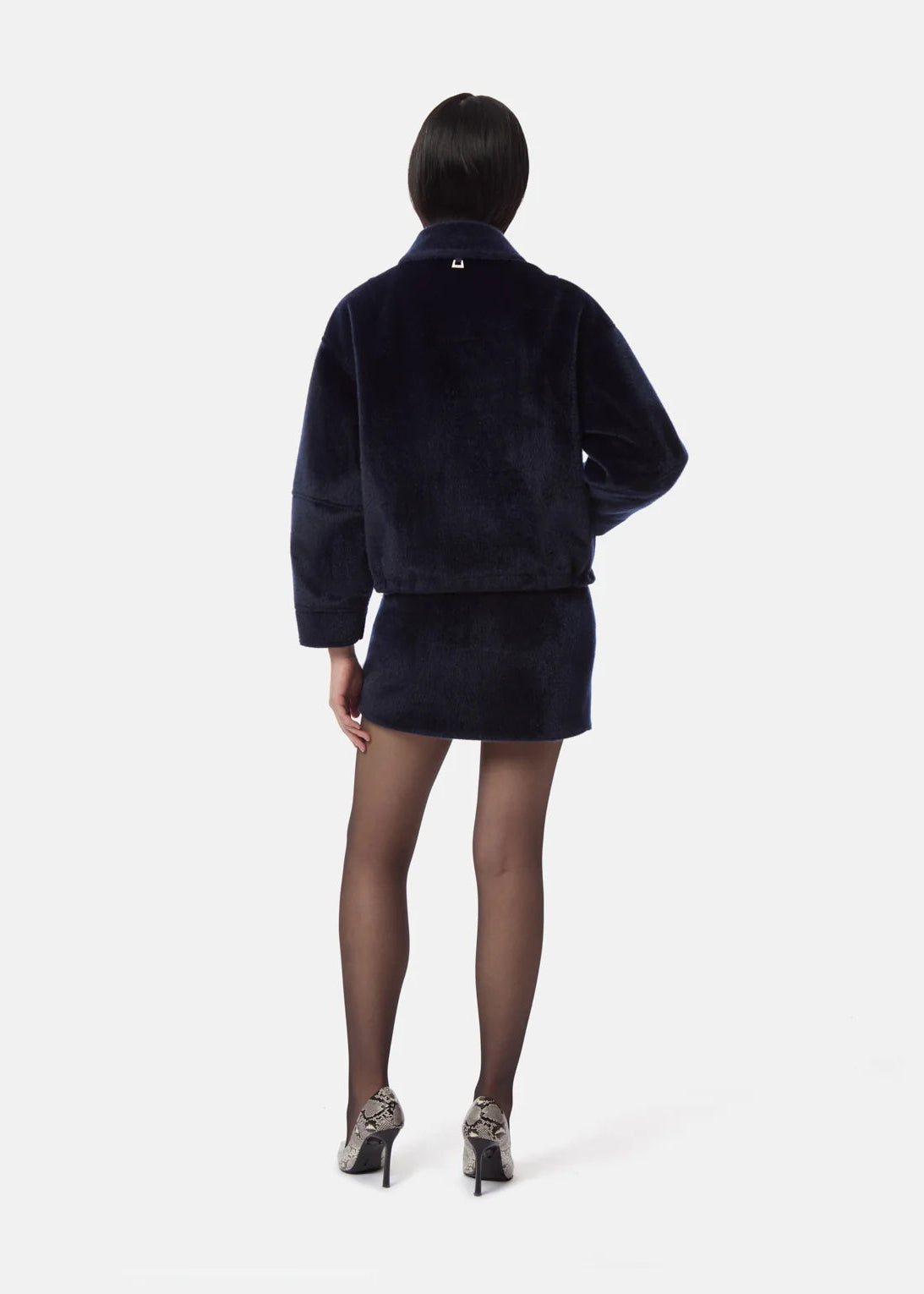 A model viewed from the back wearing a dark navy blue cropped jacket and matching mini skirt, paired with sheer black tights and patterned high heels, against a light gray studio background.