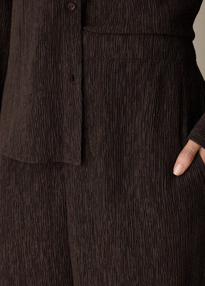 Close-up of a dark brown crinkled fabric garment, showing a buttoned section and a hand tucked into a side pocket.