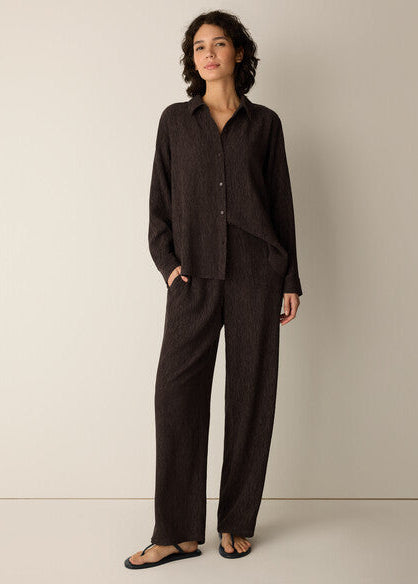 A woman modeling a dark brown textured two-piece linen set consisting of a long-sleeve button-up shirt and matching wide-leg trousers, paired with black flip-flops against a neutral beige studio background.