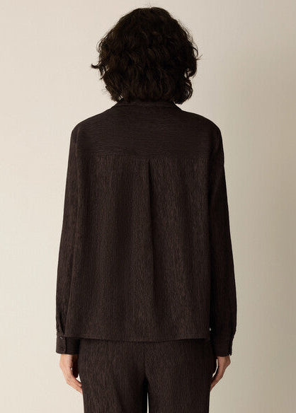 A person shown from the back wearing a dark brown textured long-sleeve shirt and matching dark pants against a plain light beige studio background.