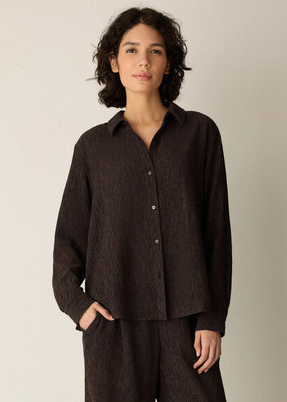 A woman wearing a dark brown textured button-up shirt and matching dark pants, standing against a plain light beige studio background.