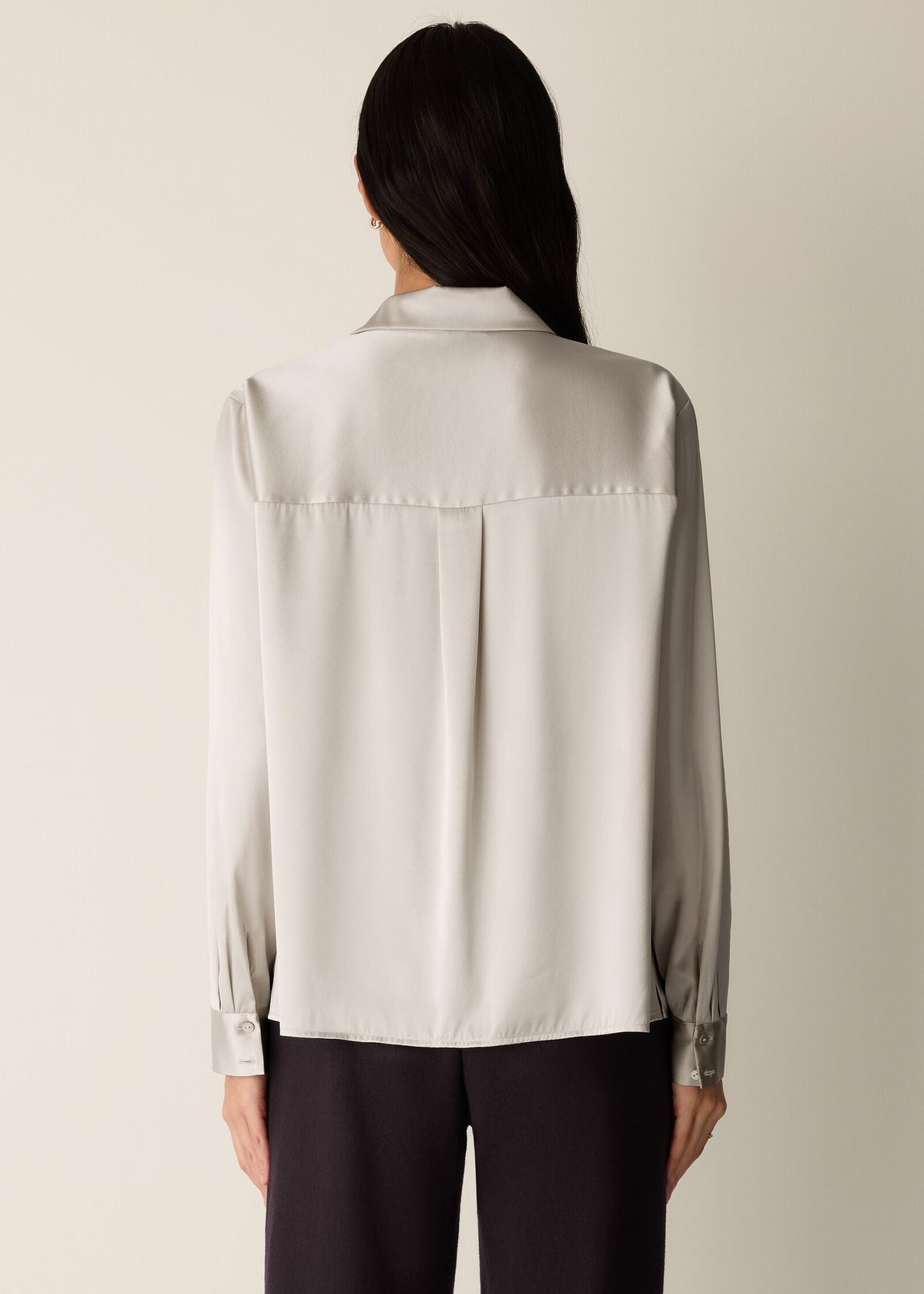 A woman shown from the back wearing a light beige or off-white long-sleeve blouse with a subtle sheen and dark trousers, set against a plain light beige studio background.