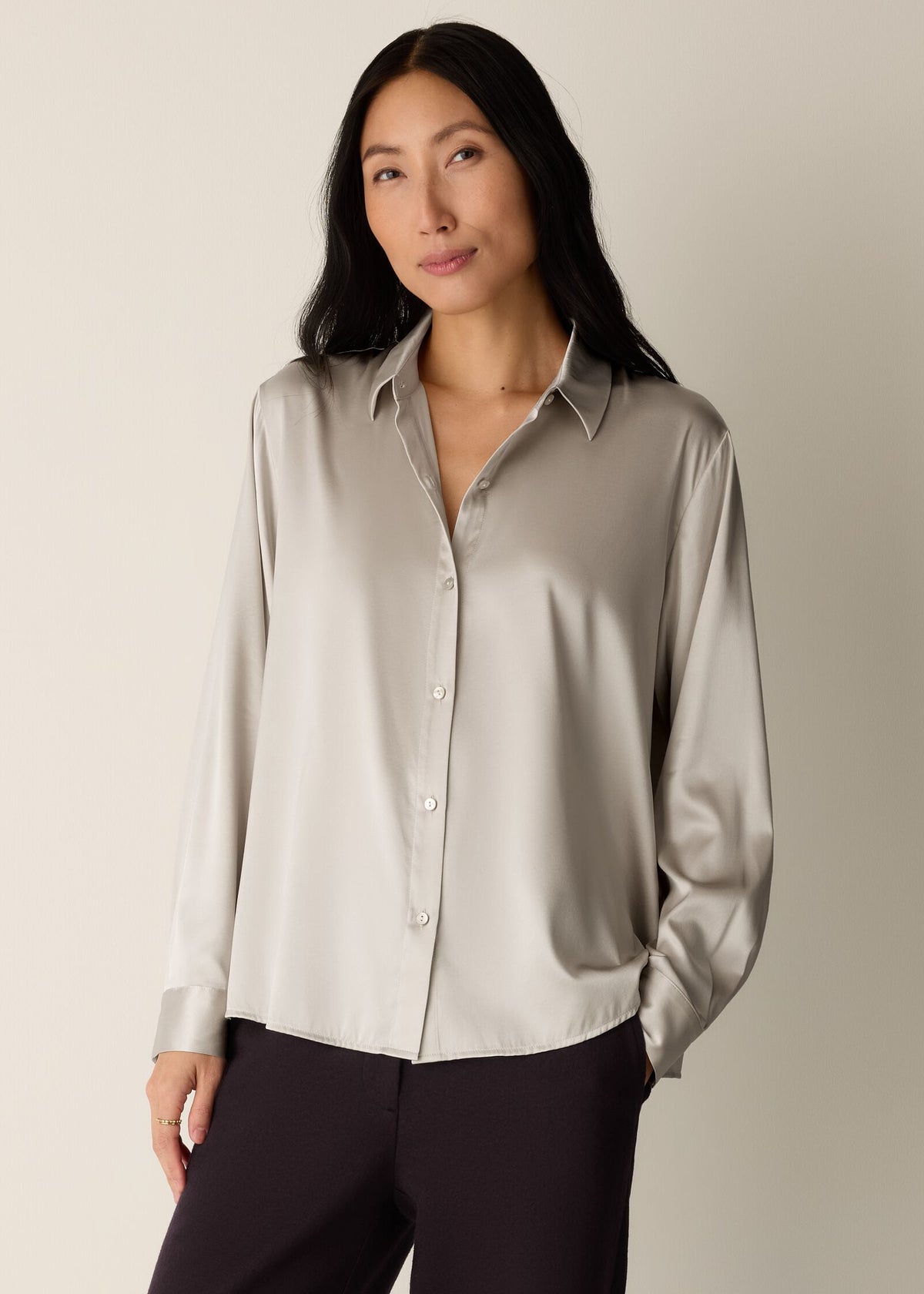 A woman modeling a long-sleeve satin button-up shirt in a light taupe-beige color, paired with dark trousers, against a plain light beige studio background.