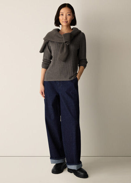 A model stands against a plain light beige studio background wearing a dark gray ribbed knit sweater with a large folded collar detail, dark blue wide-leg jeans with cuffed hems, and black leather shoes.