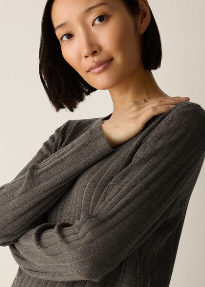 A woman with a short dark bob hairstyle wearing a dark gray ribbed knit sweater, posing with her hand on her shoulder against a plain light beige background.