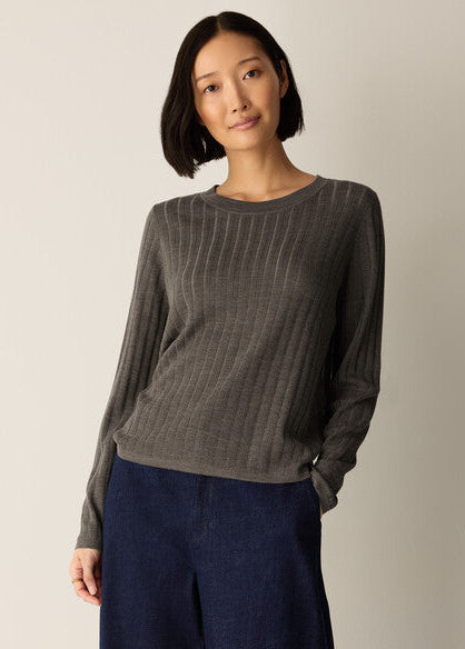 A woman wearing a dark gray ribbed long-sleeve knit top and dark blue wide-leg pants, standing against a plain light beige studio background.