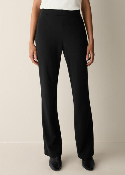 A model from the waist down wearing black wide-leg trousers, a white top, and black pointed shoes, standing against a plain beige studio background.