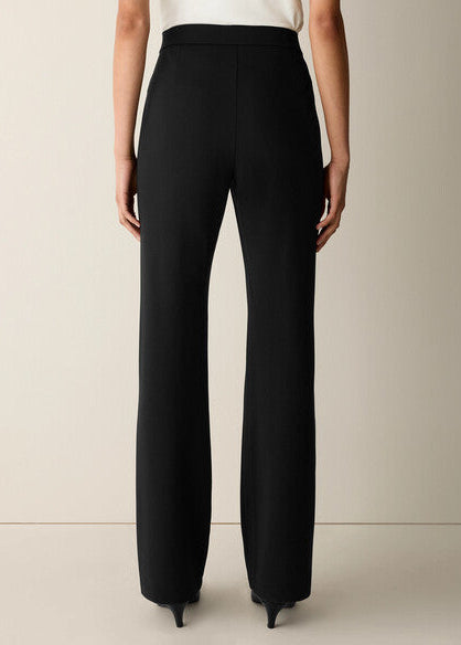A model shown from the back wearing black wide-leg trousers and black pointed-toe heels, paired with a white top, against a neutral beige studio background.