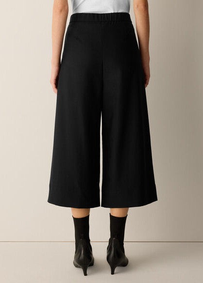 A model from the back wearing black wide-leg cropped pants, a white top, and black ankle boots against a neutral beige studio background.