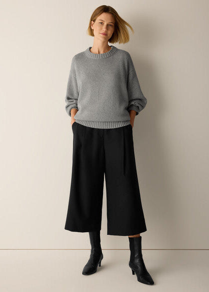 A woman stands against a plain beige studio wall wearing a light gray knit sweater, wide-leg black pants, and black ankle boots.