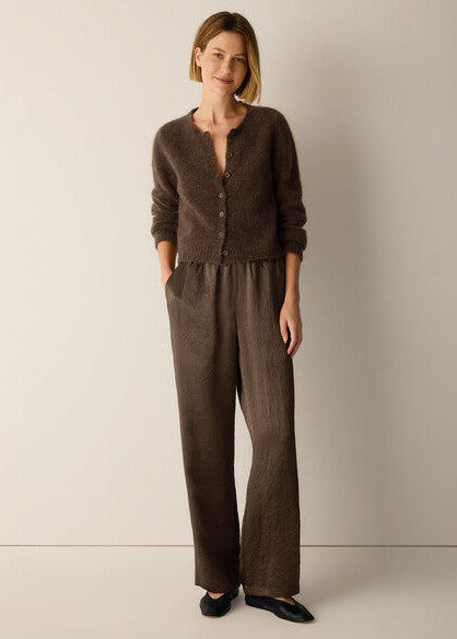 A woman stands against a plain light beige studio wall wearing a matching brown knit cardigan and wide-leg trousers set, with black flats.