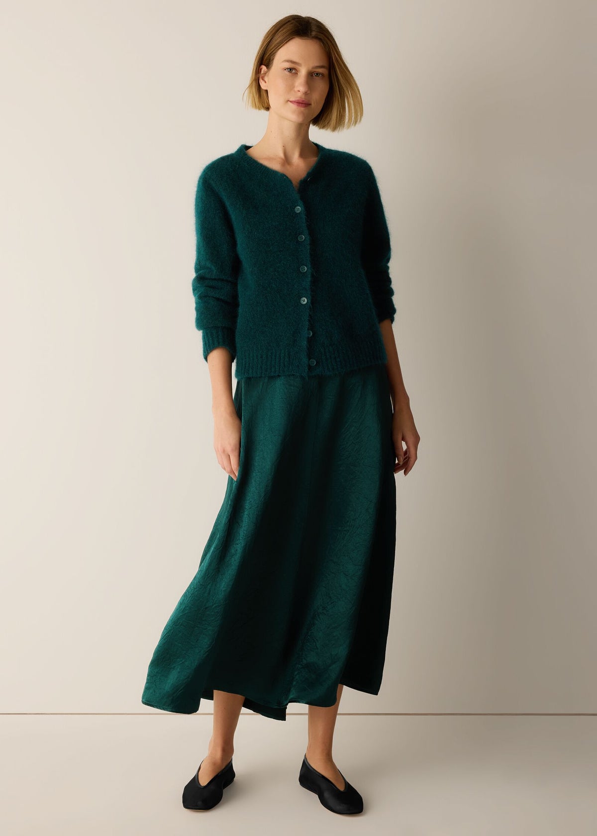 A woman modeling a dark teal green knit cardigan and matching midi skirt set, paired with black flats, against a plain beige studio background.