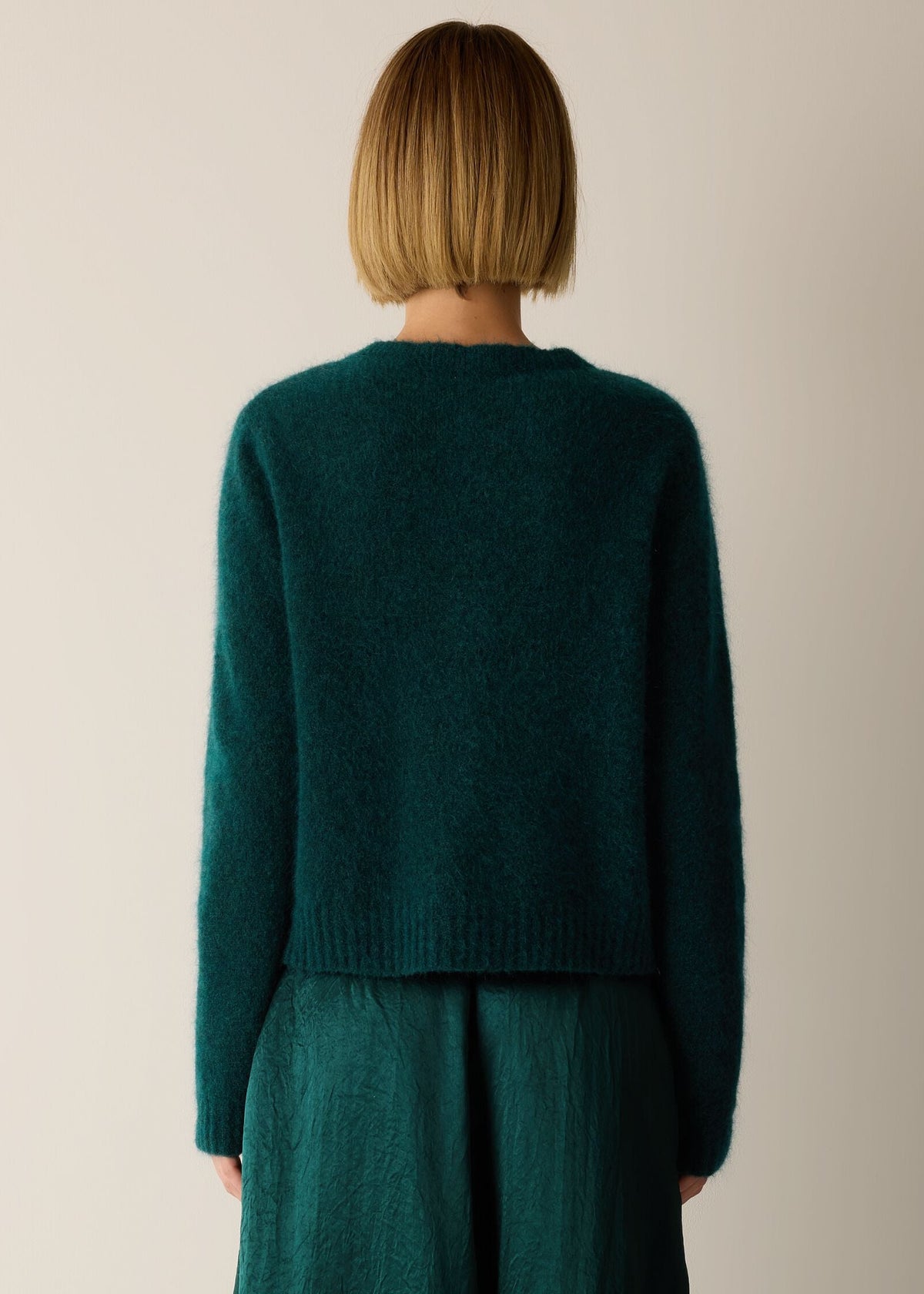 A person shown from the back wearing a dark teal-green fuzzy knit sweater and matching dark green skirt against a plain light beige studio background.