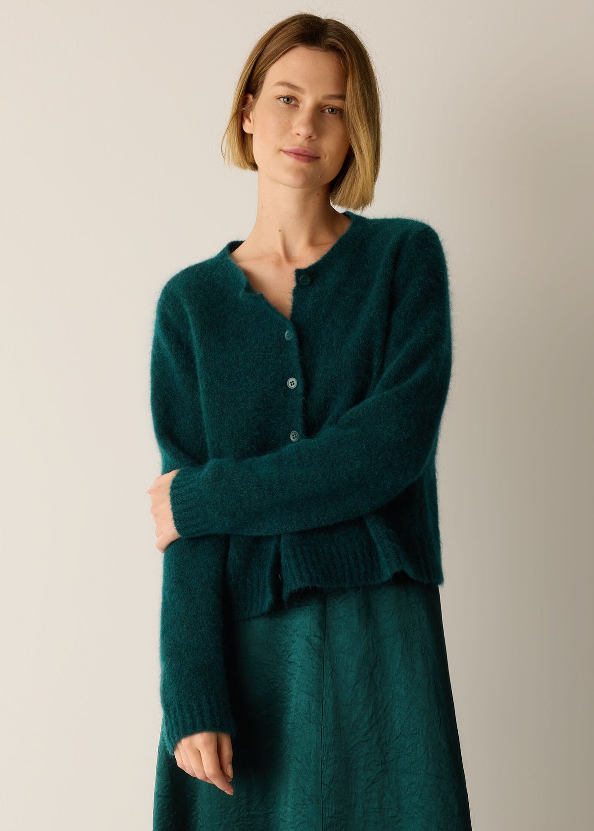 A woman wearing a dark teal green knit cardigan with a buttoned neckline over a matching knit dress, standing against a plain light beige studio background.