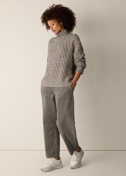 A woman modeling a gray cable-knit mock-neck sweater and matching gray wide-leg trousers with white sneakers, standing against a plain light beige studio background.