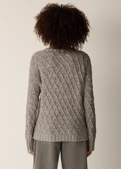 A person shown from the back wearing a gray cable-knit sweater and matching gray pants against a plain light beige studio background.