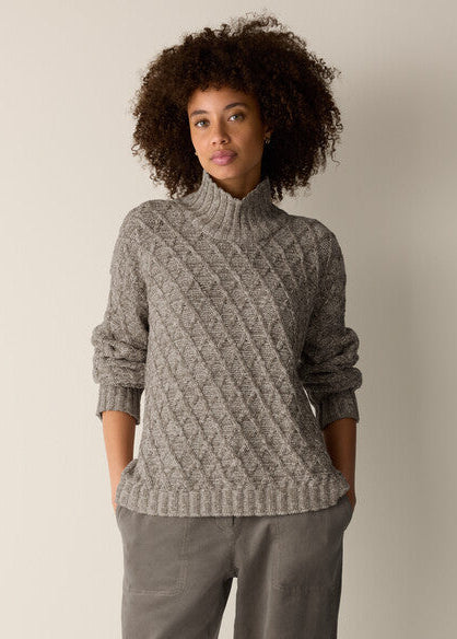 A woman with curly dark hair wearing a gray cable-knit mock neck sweater and matching gray pants, standing against a plain light beige studio background.