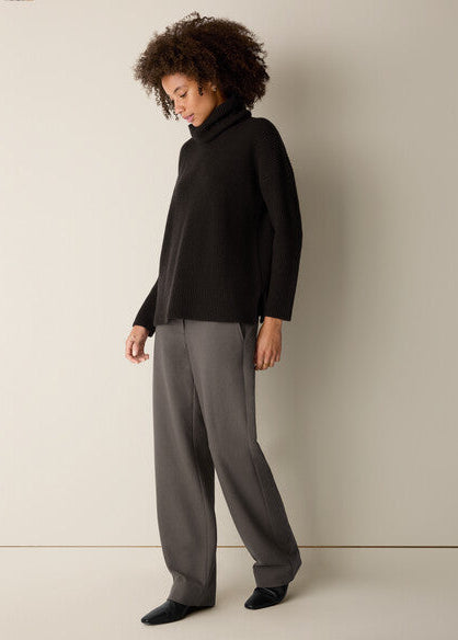 A model with curly dark hair is shown from the side, wearing a black long-sleeve mock neck top and loose, wide-leg gray trousers against a plain beige studio background.