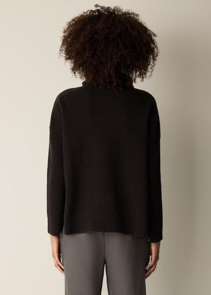 A person shown from the back wearing a black ribbed knit turtleneck sweater and gray trousers against a plain light beige studio background.