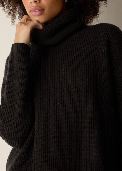 Close-up of a person wearing a black ribbed turtleneck sweater, with the hand near the neck and the collar pulled high. The background is a plain light neutral wall.