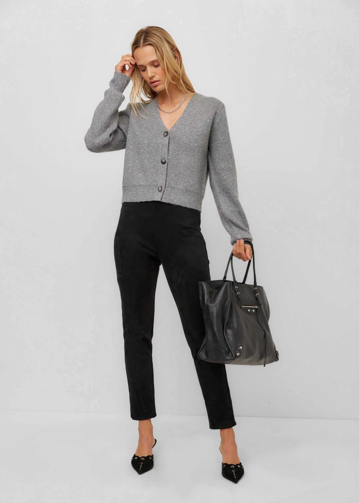 A woman modeling a light gray long-sleeve button-up cardigan, black cropped trousers, and black pointed-toe heels, while holding a black leather tote bag against a light gray studio background.