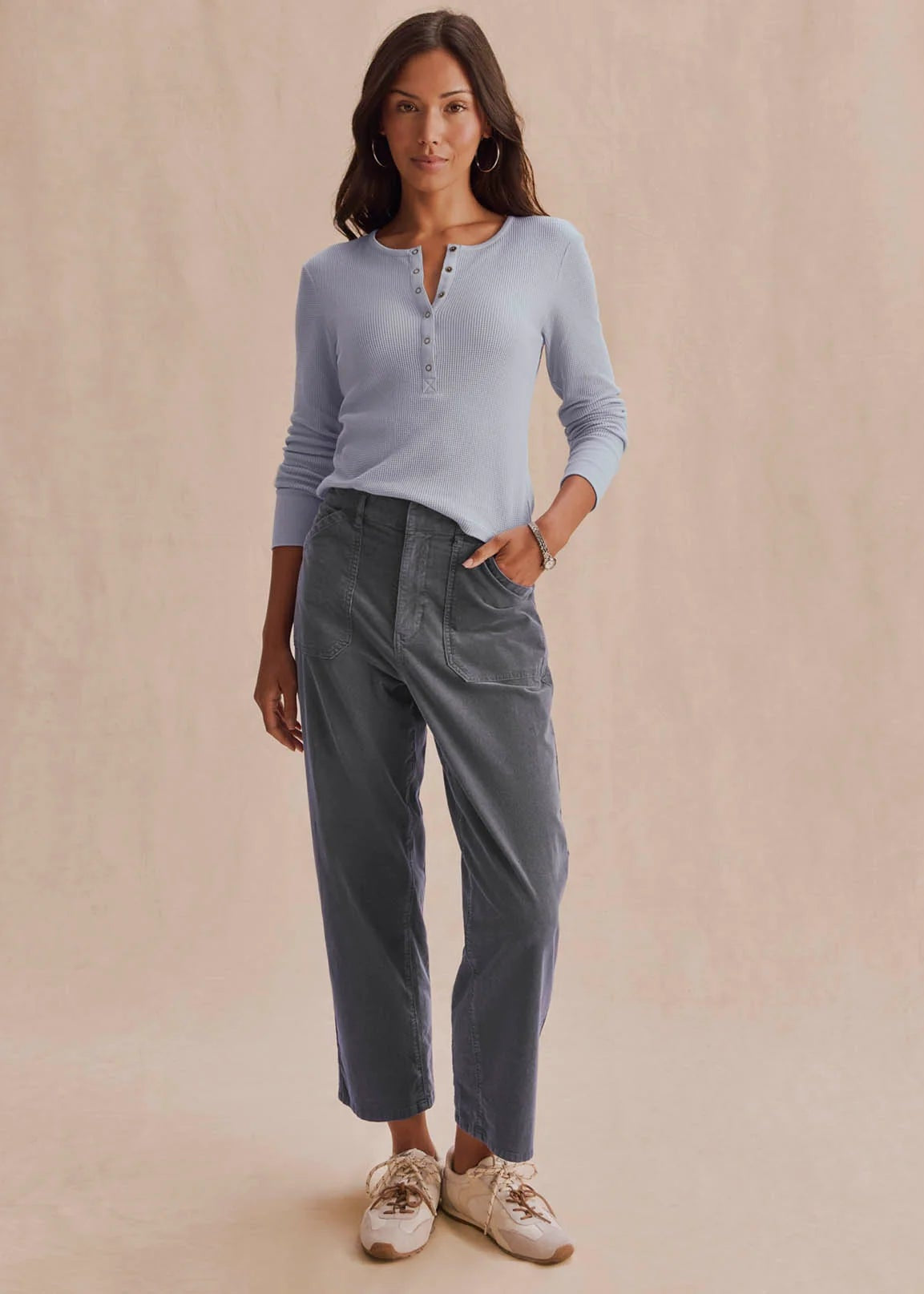 A woman modeling a light blue long-sleeve henley top and dark gray cropped pants with beige sneakers, standing against a warm beige textured studio backdrop.