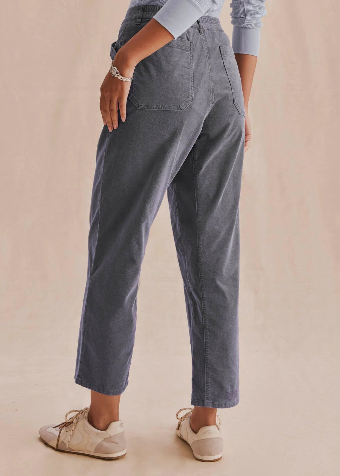 A person shown from the back wearing slate blue cropped pants, a light blue long-sleeve top, and beige sneakers against a plain beige studio background.