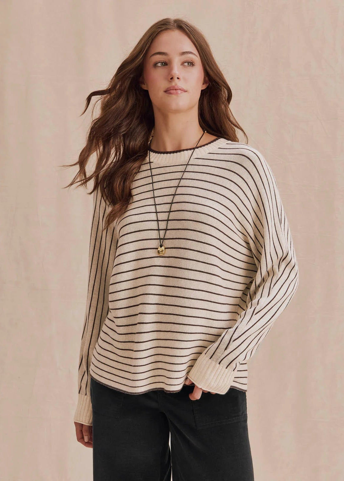A woman wearing a cream and black horizontally striped knit sweater with black pants, standing against a neutral beige backdrop.