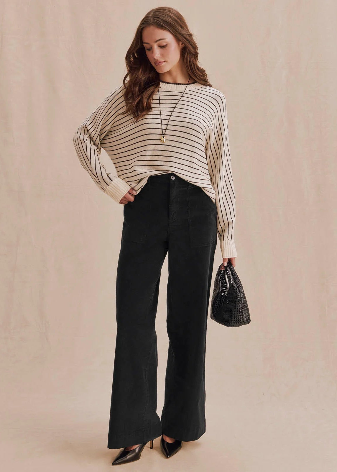 A woman models a cream and black horizontally striped knit sweater paired with black wide-leg pants, black heels, and a small black woven handbag, set against a warm beige textured backdrop.