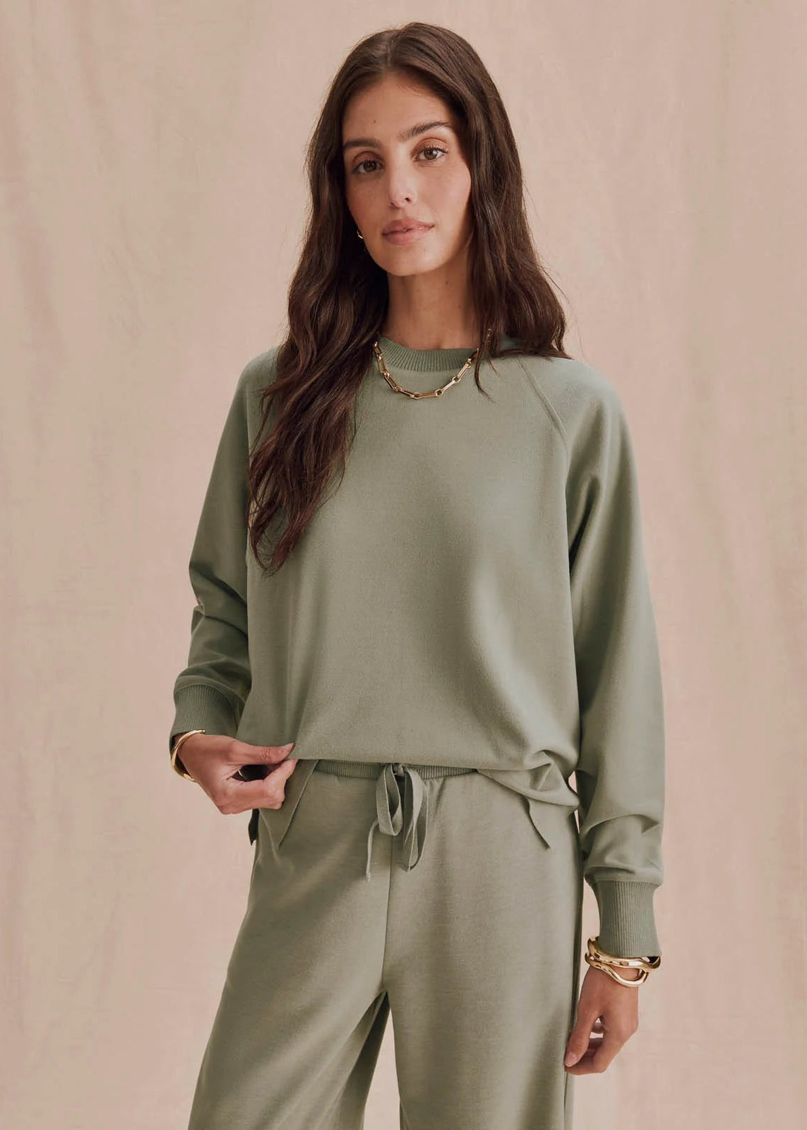 A woman modeling a matching sage green lounge set consisting of a long-sleeve crewneck sweatshirt and drawstring pants, set against a neutral beige textured background.