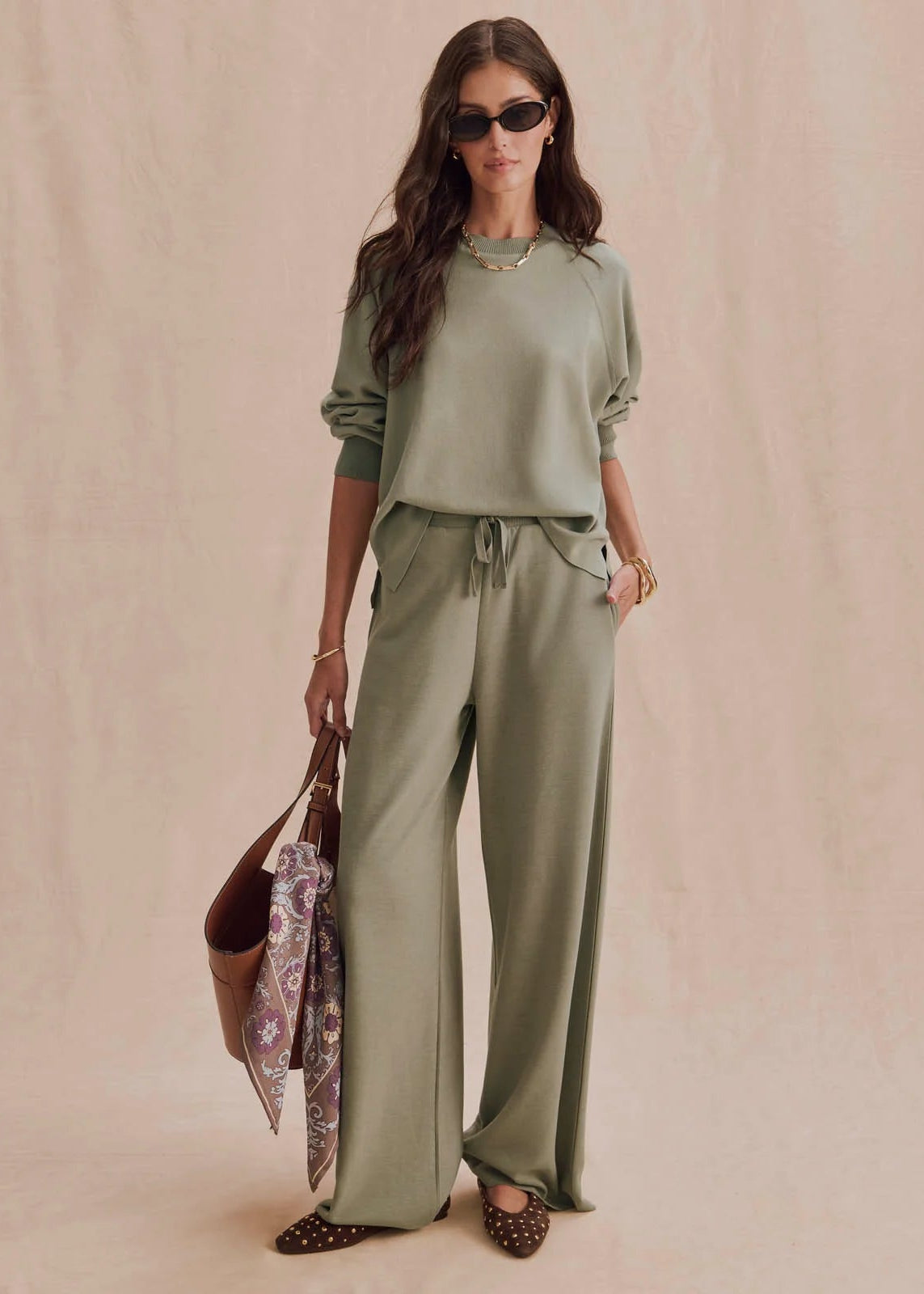 A woman models a matching sage green lounge set consisting of a long-sleeve top and wide-leg drawstring pants, styled with sunglasses, a patterned scarf, and brown pointed flats against a soft beige backdrop.