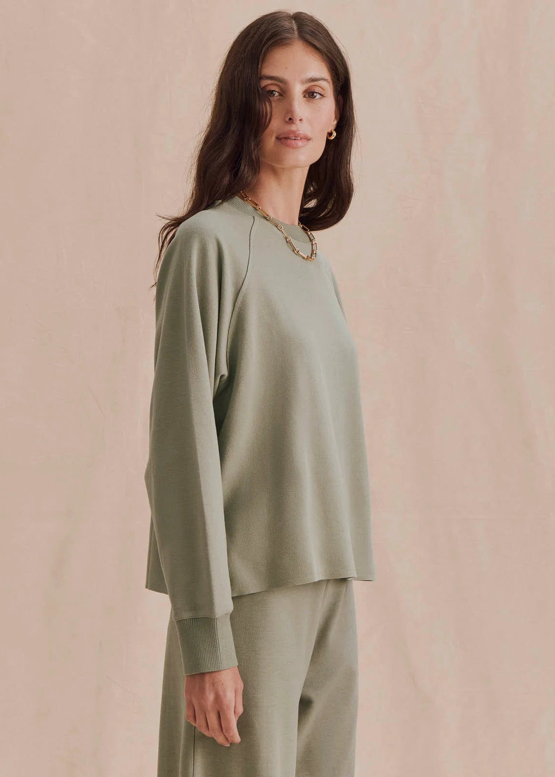 A woman modeling a sage green long-sleeve knit top and matching wide-leg pants against a soft beige textured backdrop.