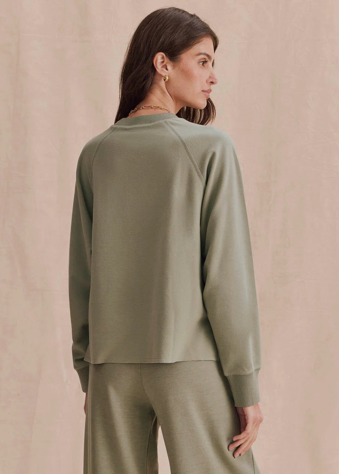 A woman shown from the back wearing a sage green long-sleeve top and matching wide-leg pants against a soft beige backdrop.