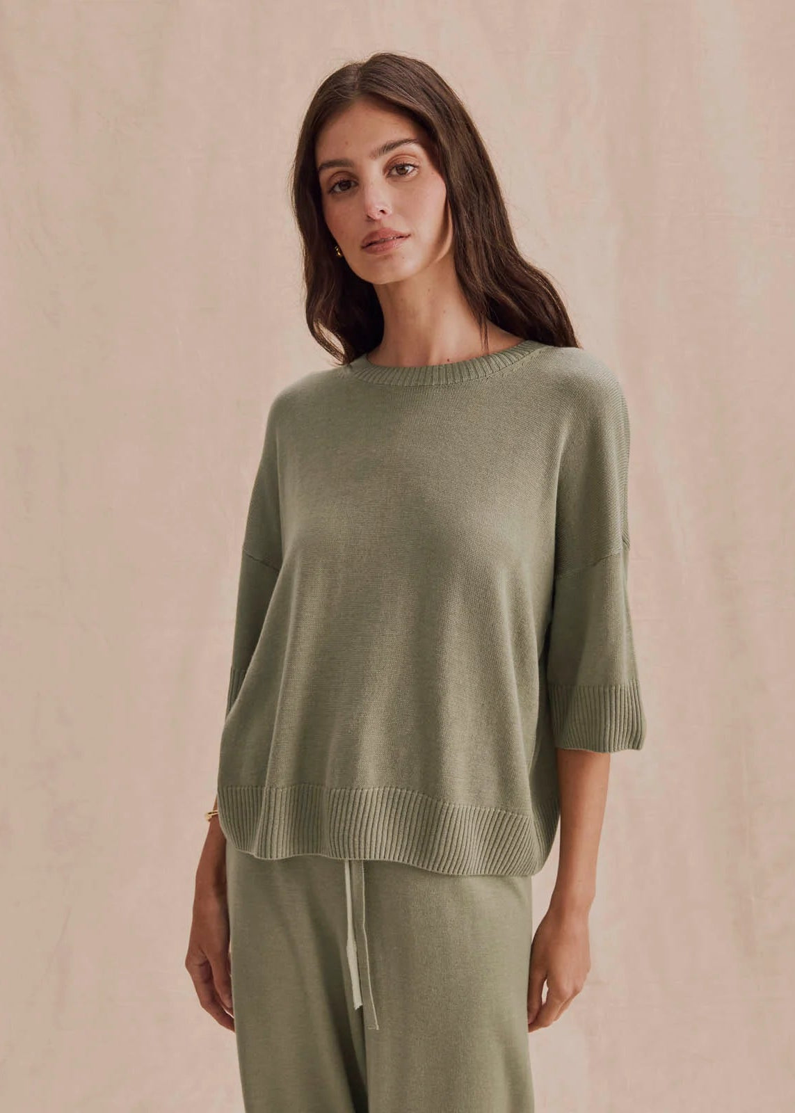 A woman modeling a light sage green knit sweater and matching knit pants against a soft beige textured backdrop.