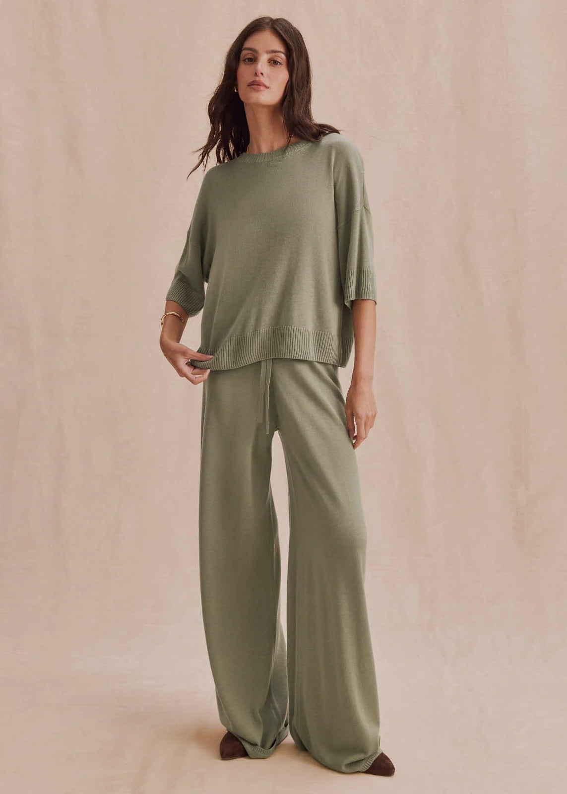 A woman modeling a matching sage green lounge set consisting of a short-sleeve crewneck top and wide-leg pants, standing against a textured beige backdrop.