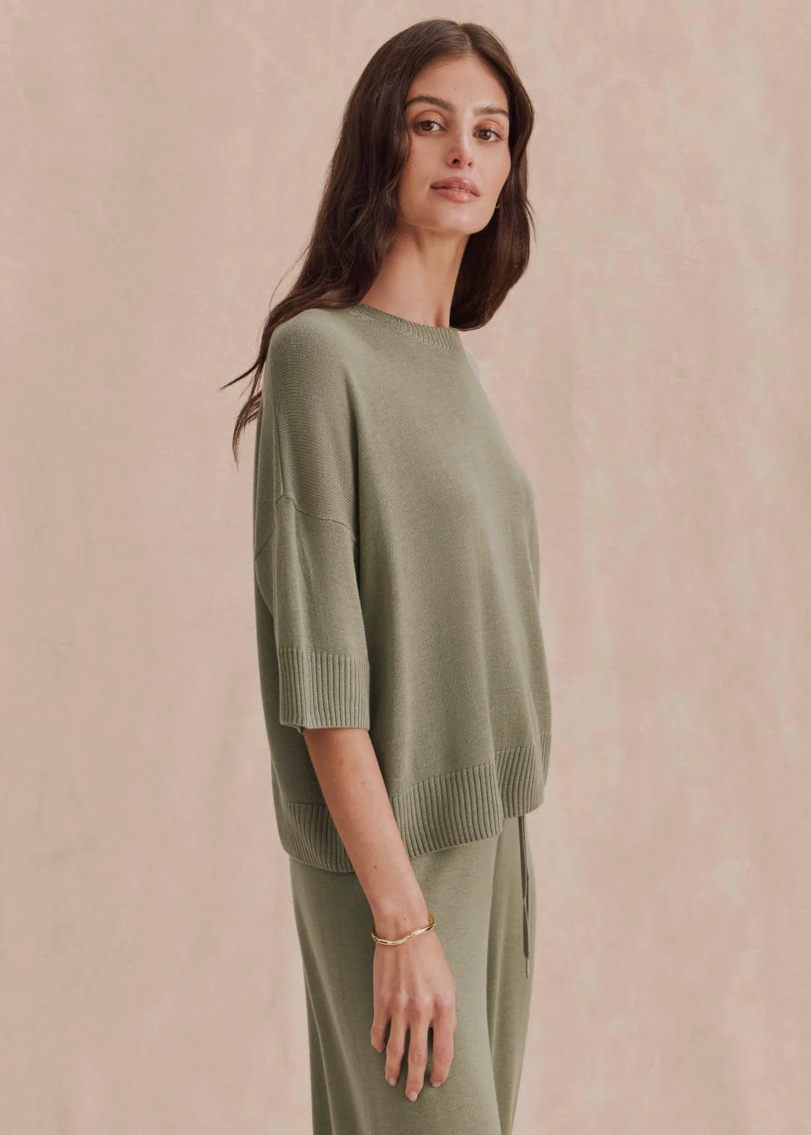 A woman modeling a light sage green knit sweater and matching knit pants against a soft beige textured background.
