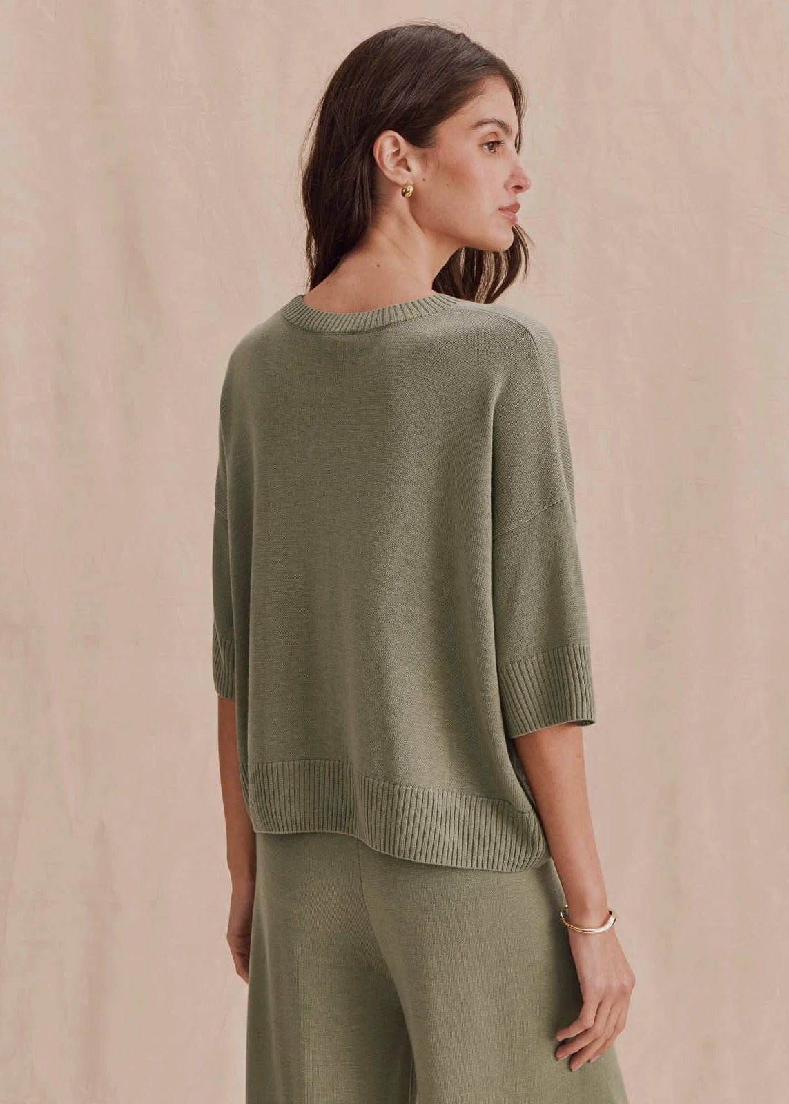 A woman wearing a muted sage green knit sweater and matching knit pants, photographed from the back against a soft beige studio backdrop.