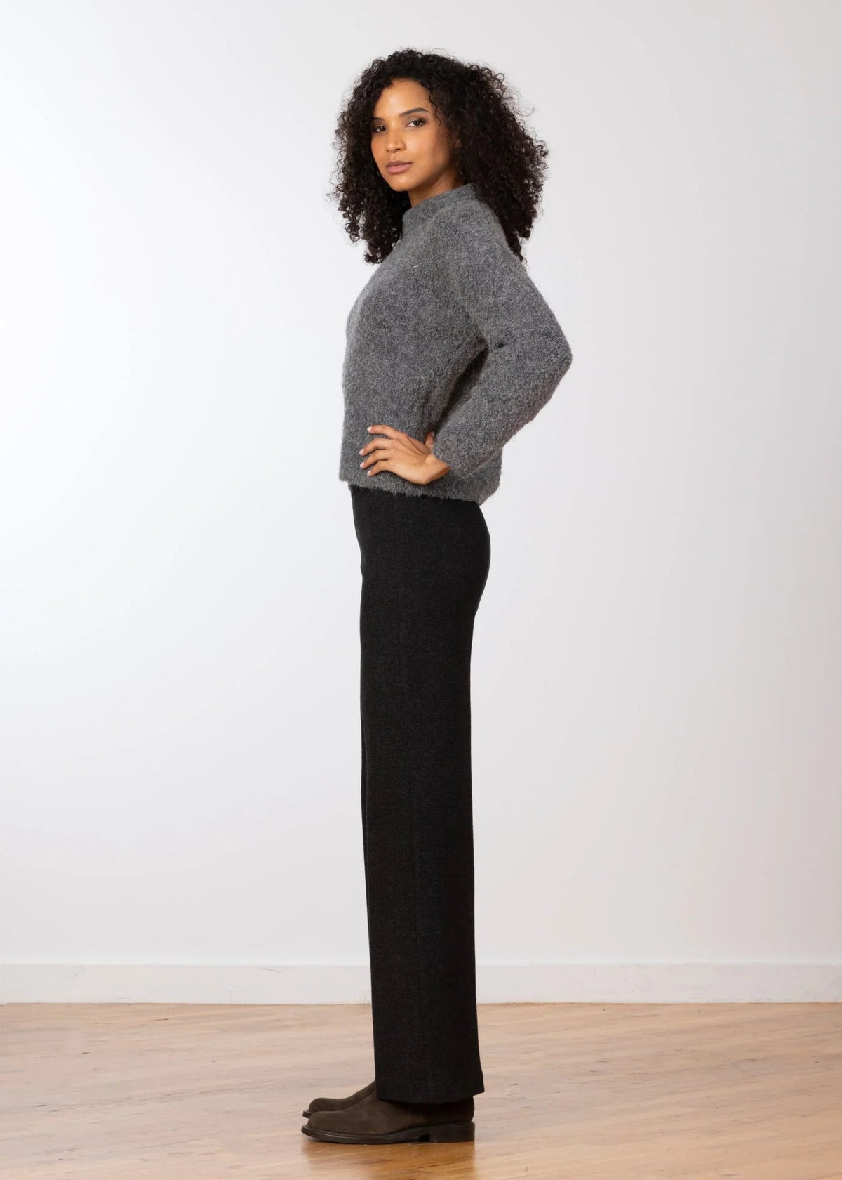 A woman stands in profile wearing a fuzzy gray mock-neck sweater and black wide-leg pants against a plain white wall and light wood floor.
