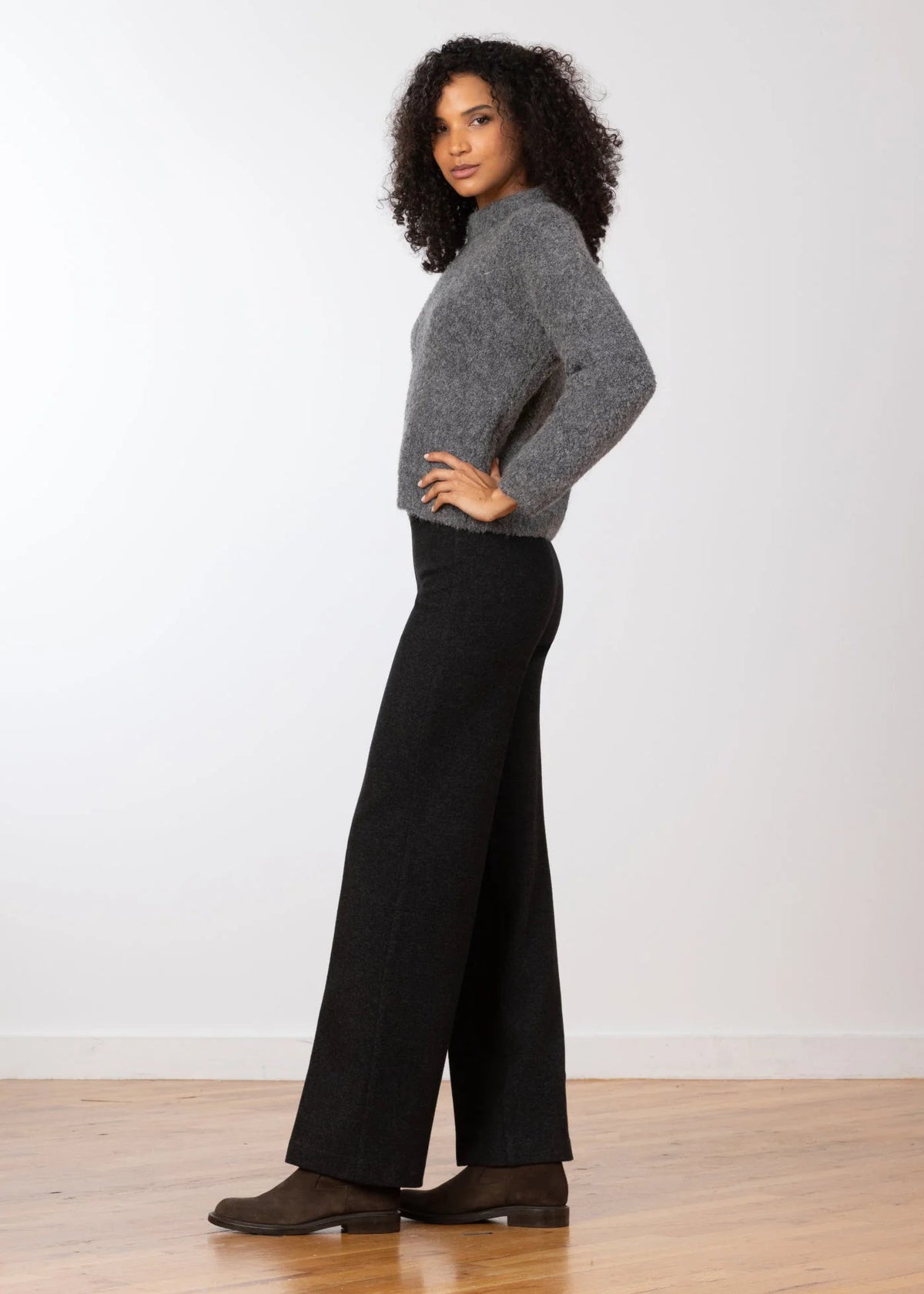 A woman stands in profile wearing a gray knit mock-neck sweater and black wide-leg pants, paired with dark brown shoes, against a white wall and light wood floor.
