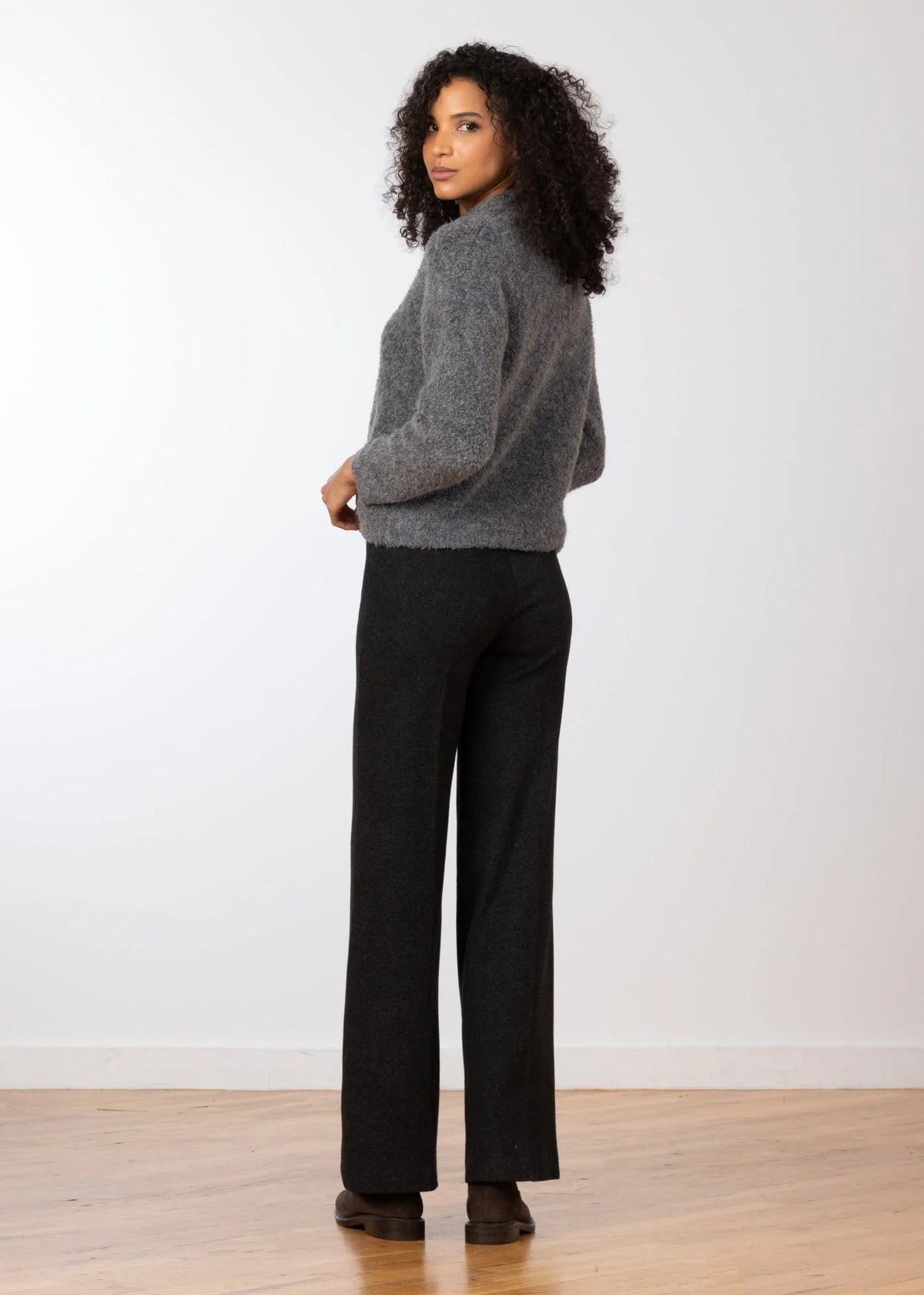 A woman stands in profile wearing a gray knit turtleneck sweater and black wide-leg trousers against a plain white wall and light wood floor.