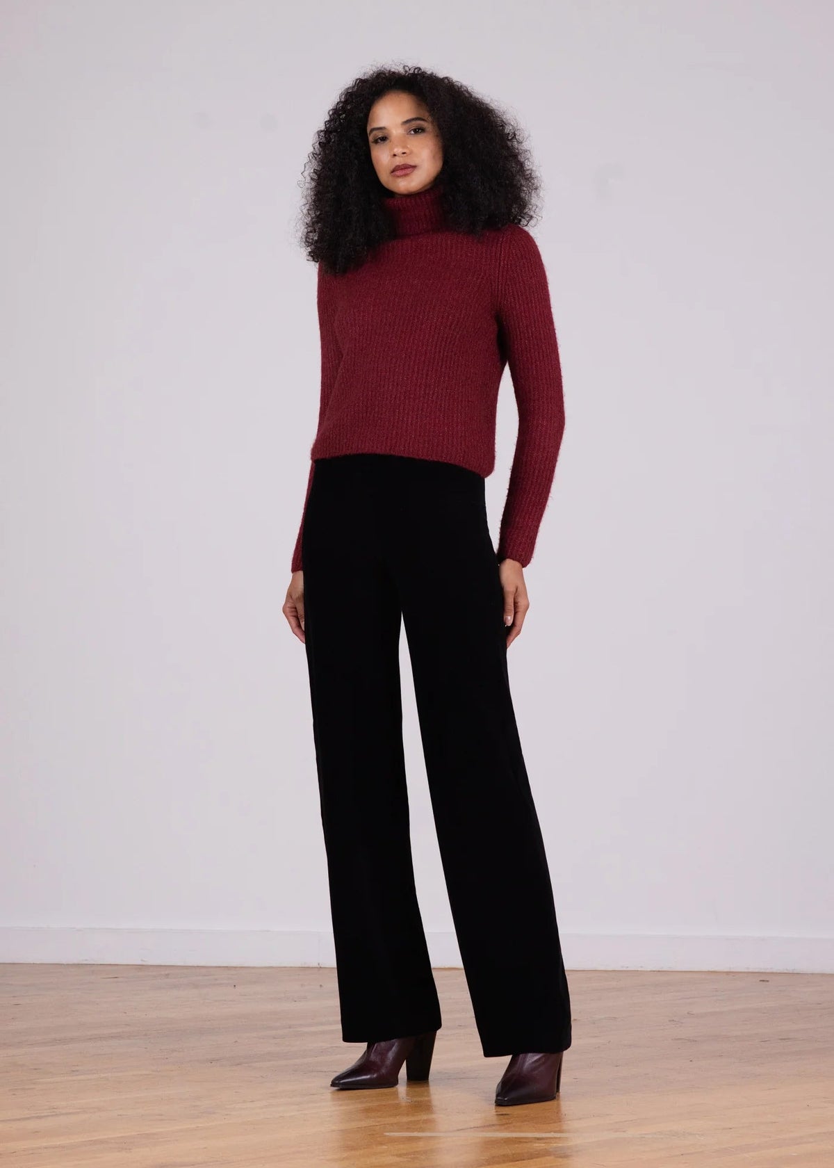 A woman stands in a studio wearing a burgundy ribbed turtleneck sweater and black wide-leg trousers, paired with dark brown pointed-toe heels, against a light gray wall and wood floor.
