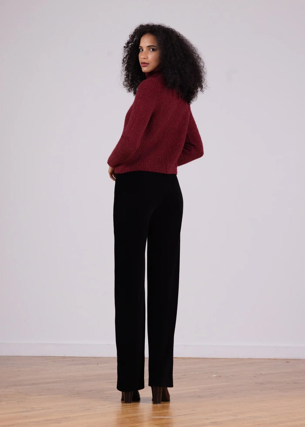 A woman stands in profile wearing a ribbed burgundy cropped sweater and black wide-leg trousers, standing on a light wood floor against a plain white wall.
