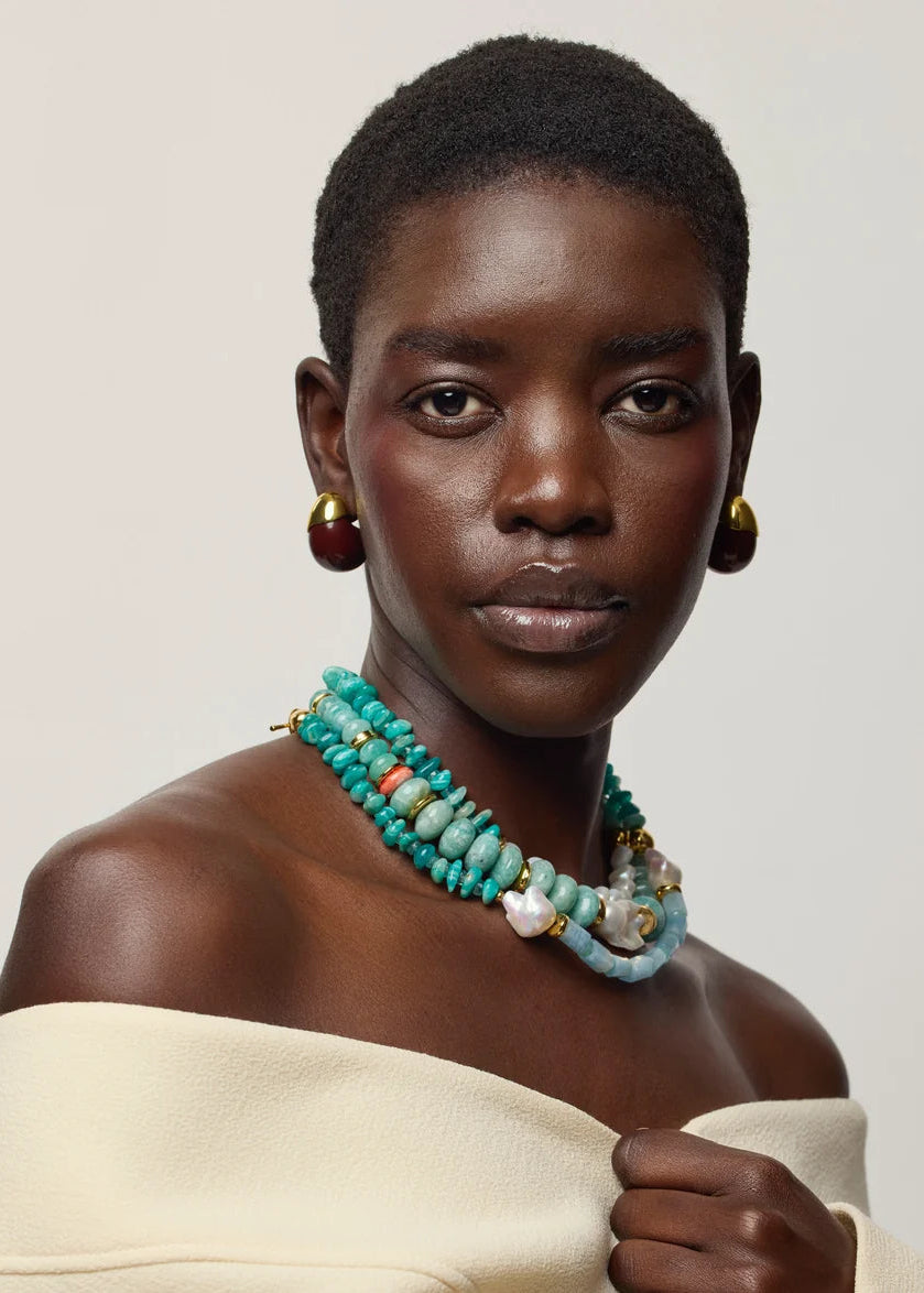 A close-up portrait of a model wearing a multi-strand turquoise beaded necklace with white and orange accents, gold hoop earrings, and a cream off-the-shoulder top against a light gray background.