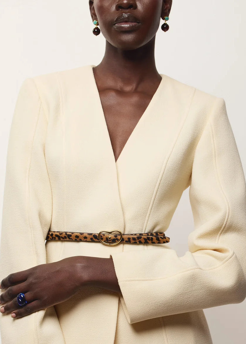 A close-up of a model wearing a cream-colored wrap jacket with a leopard-print belt featuring a gold-tone double-G buckle, styled with dark green drop earrings and a blue ring.