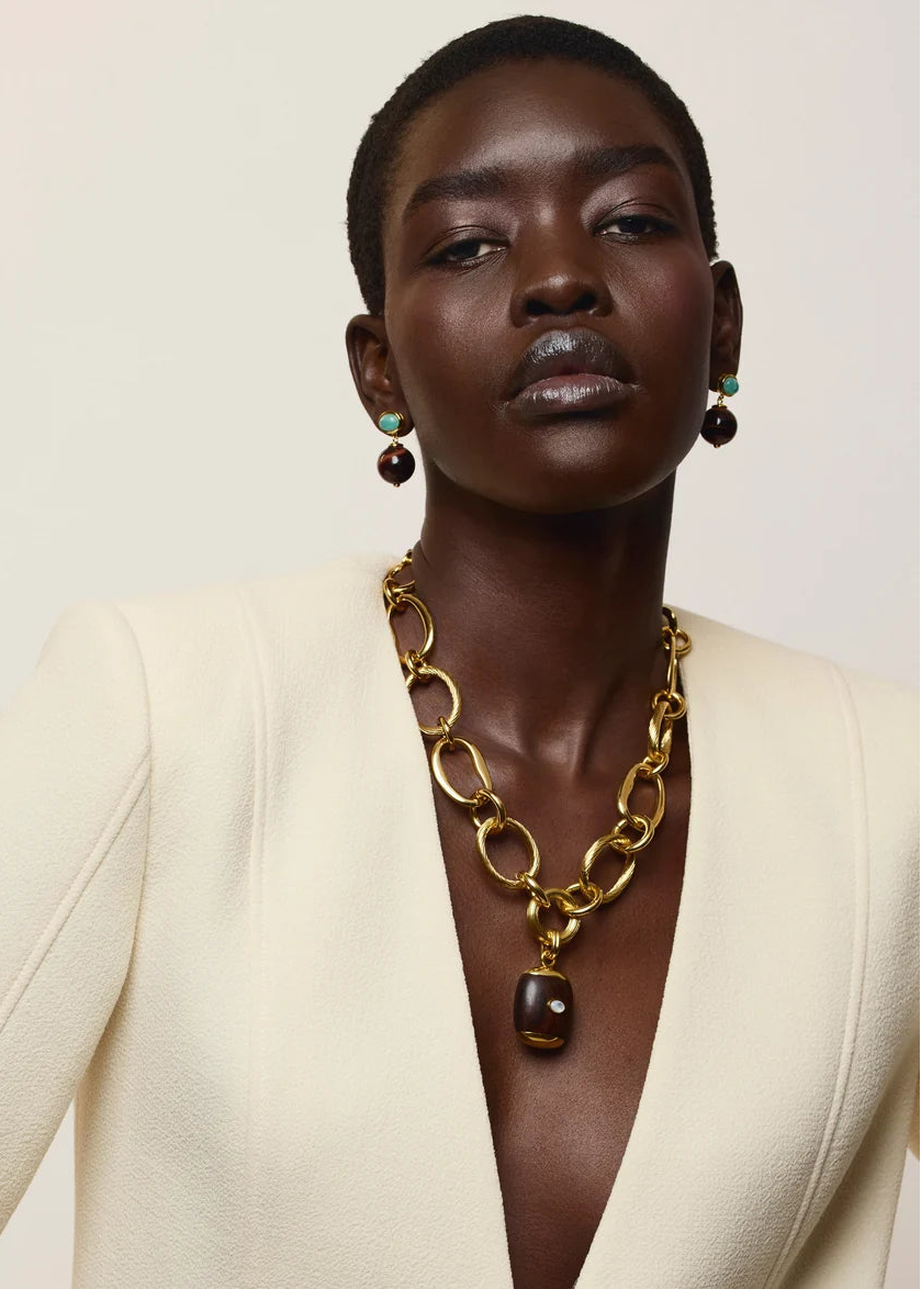 A close-up portrait of a model wearing a chunky gold chain necklace with a dark brown pendant, styled with matching drop earrings and a cream-colored blazer against a light beige background.