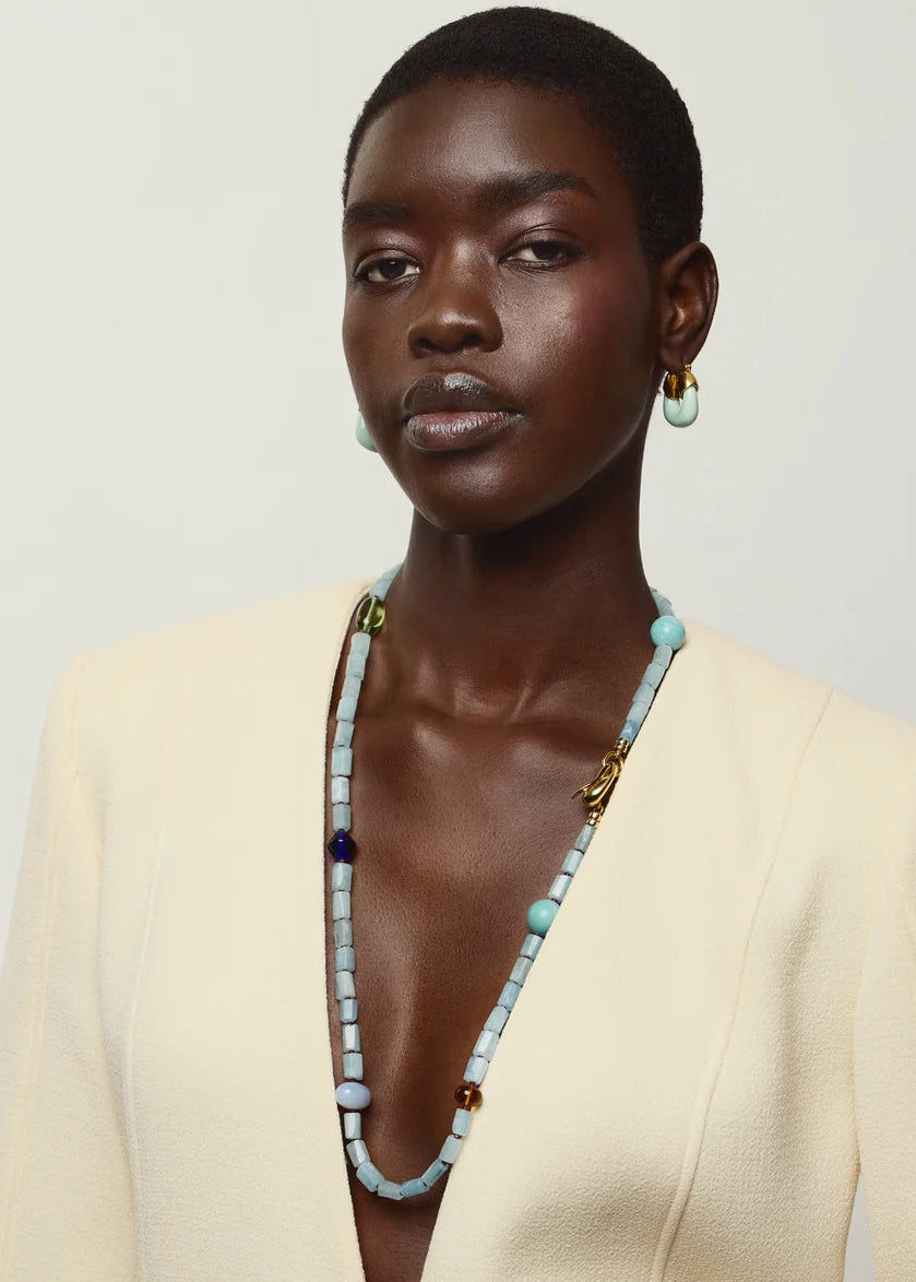 A close-up portrait of a model wearing a cream-colored blazer, a long light blue beaded necklace with gold accents, and matching light blue drop earrings against a plain light gray background.