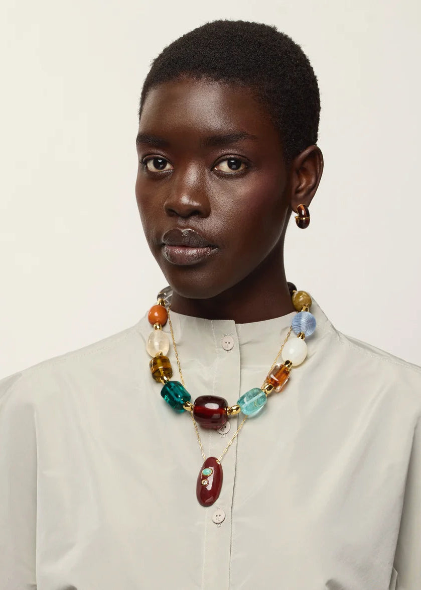 A model with short dark hair is wearing a cream-colored high-neck button-up shirt and a chunky, multi-colored beaded necklace with a red pendant, set against a plain off-white background.