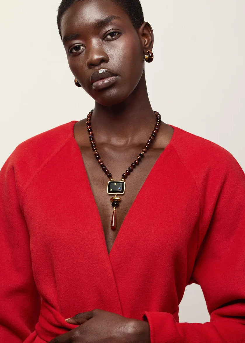 A model wearing a vibrant red knit wrap sweater and a dark brown beaded necklace with a gold and black rectangular pendant, set against a plain off-white background.