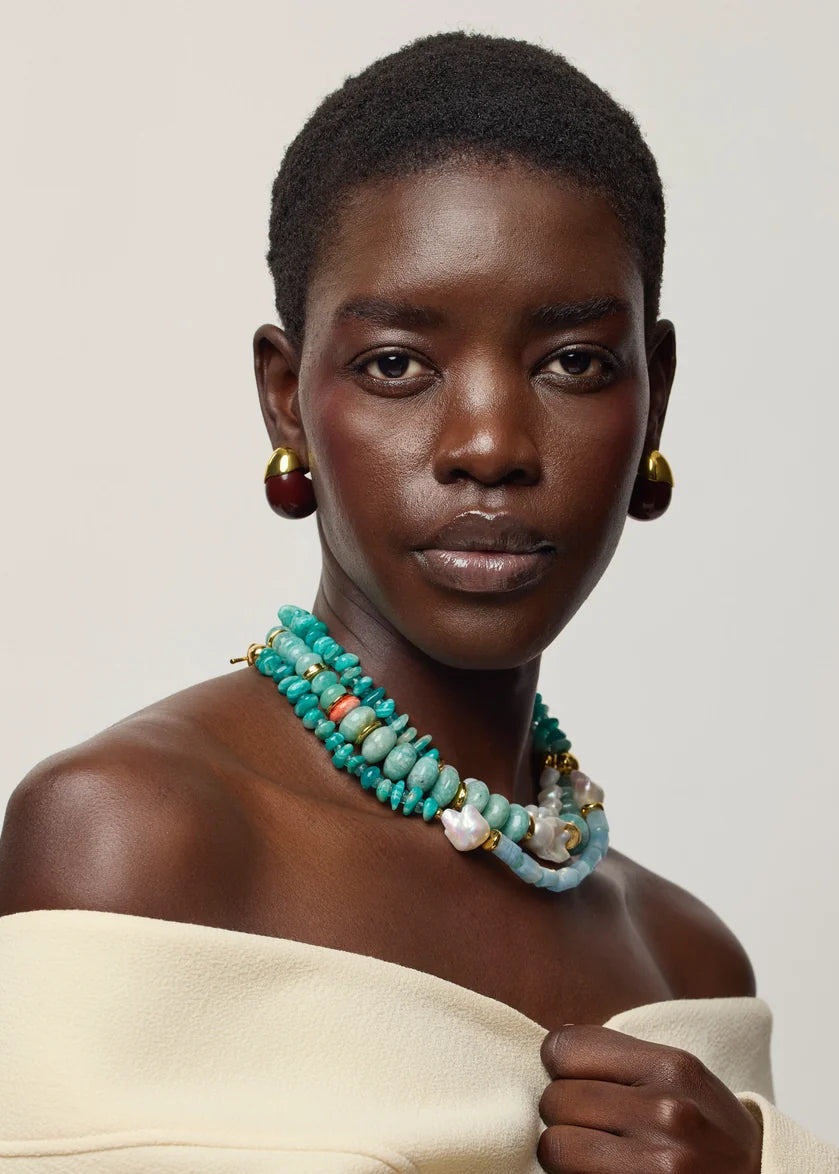 A close-up portrait of a model wearing a multi-strand turquoise beaded necklace with white and orange accents, gold hoop earrings, and a cream off-the-shoulder top against a light gray background.