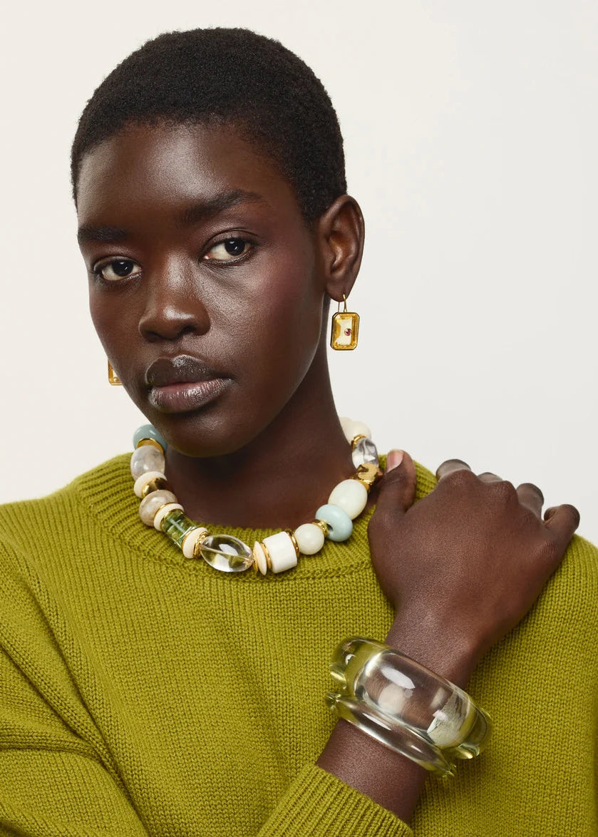A model with short dark hair is shown from the chest up, wearing a textured olive green knit sweater, a chunky beaded necklace with white, cream, and light green beads, gold-toned earrings, and a clear acrylic bangle. The background is a clean, solid off-white studio backdrop.